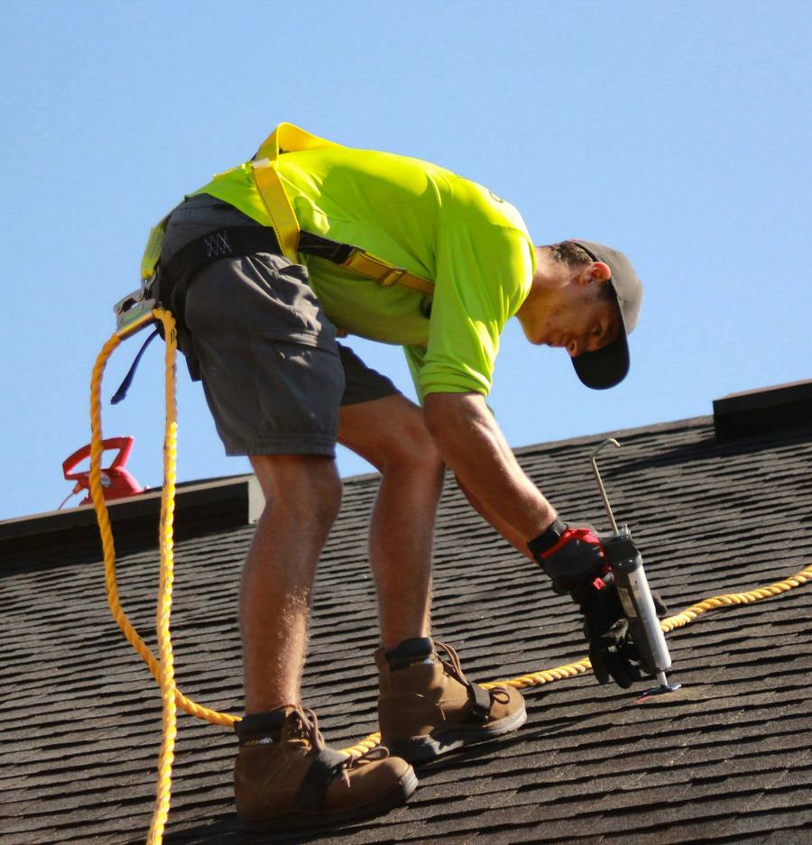 Roofer sealing a fastener on a shingle roof against a clear blue sky