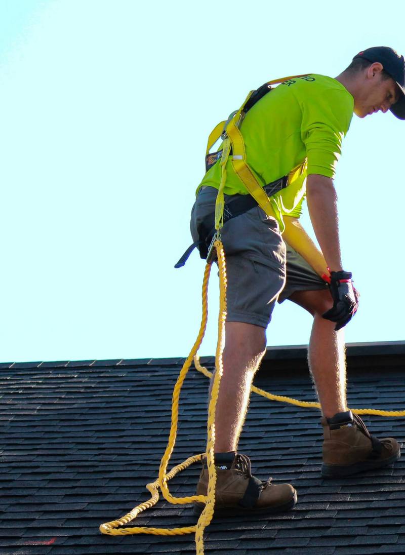 Roofer in a safety harness standing on a shingle roof against a clear sky